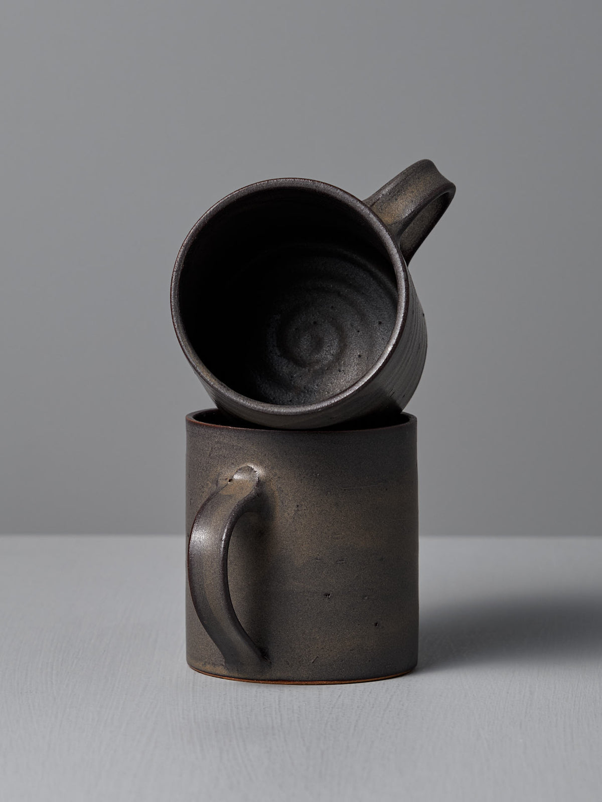Two Zoë Isaacs Coffee Cup – Dapple Brown mugs are arranged on a plain gray background—one upright and the other artfully tilted—showcasing their handmade quality and matte brown glaze.