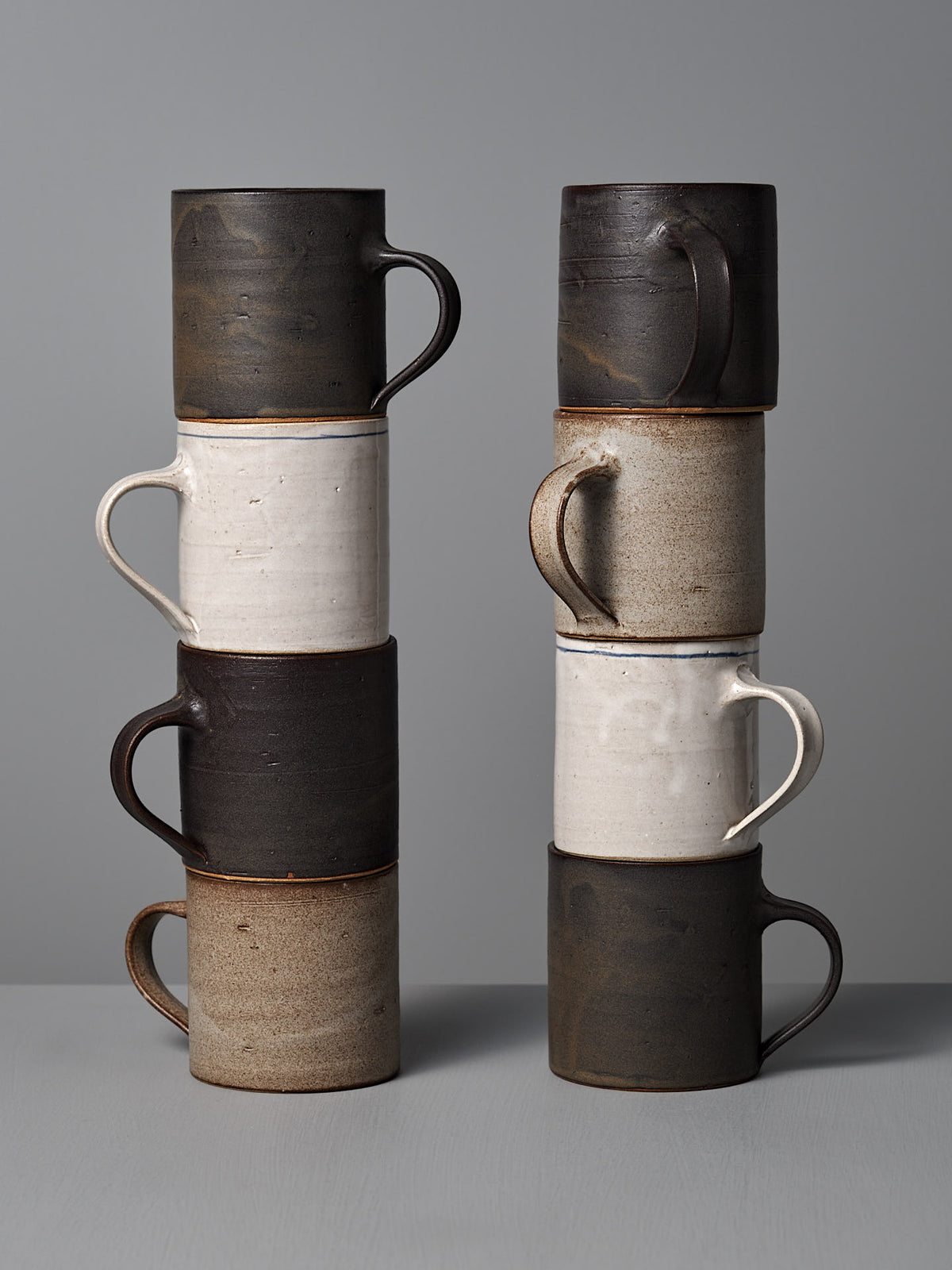 Two stacks of Zoë Isaacs Coffee Cup – Dapple Brown mugs, each with four handmade ceramic pieces featuring alternating black, white, and matte brown glazes, are displayed against a plain gray background.