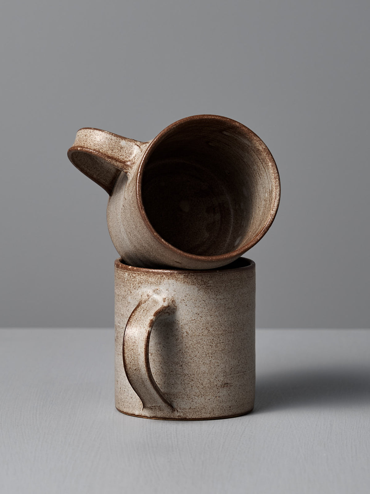Two handmade Zoë Isaacs Coffee Cups in Blue Grey are stacked—a matte glazed pair, one upright and the other sideways—set against a plain gray background.