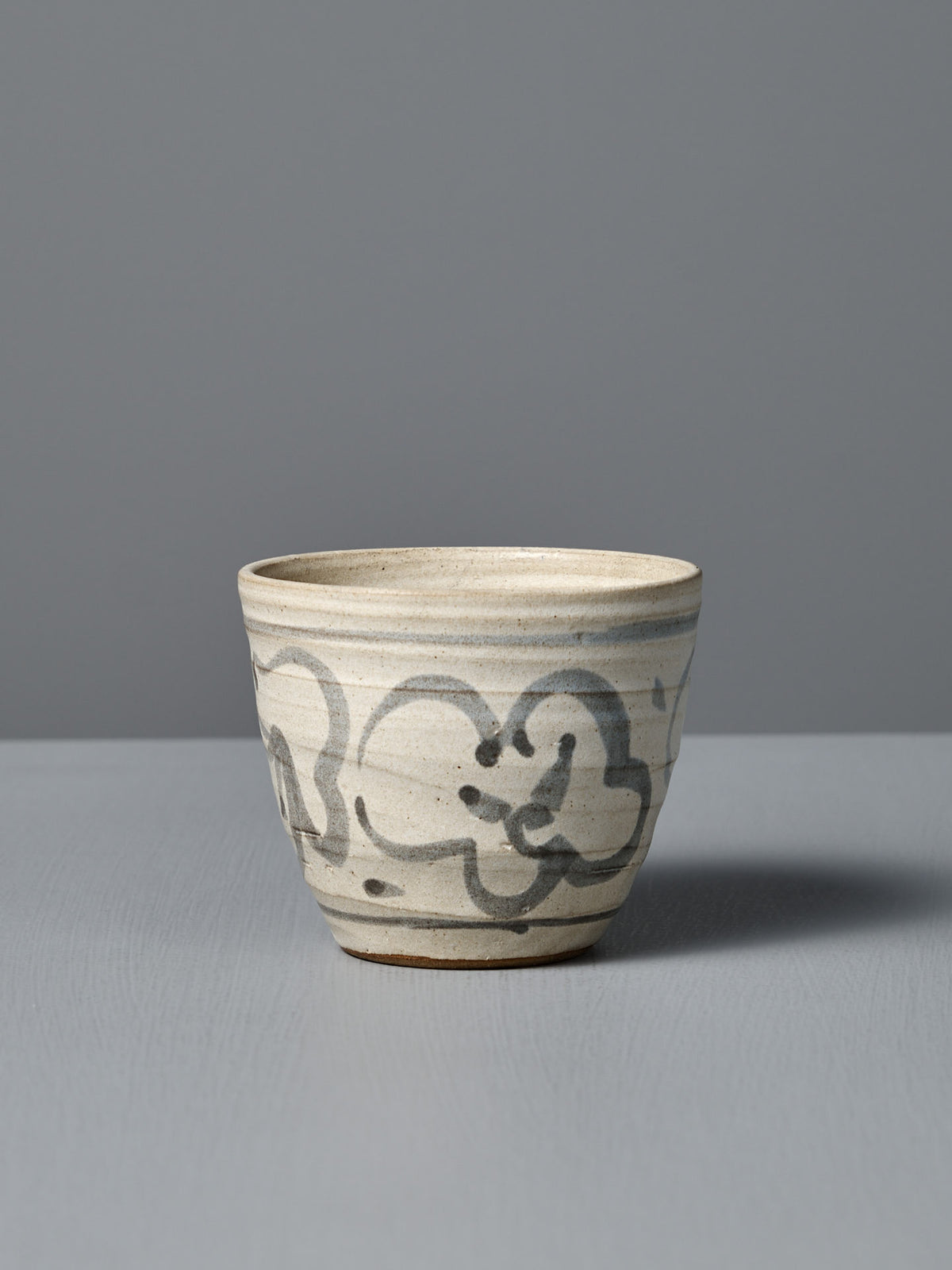 The Cup – Floral is a handmade ceramic cup featuring a beige glaze with dark abstract brushstrokes. It sits on a light gray surface against a gray background. Made in Aotearoa.