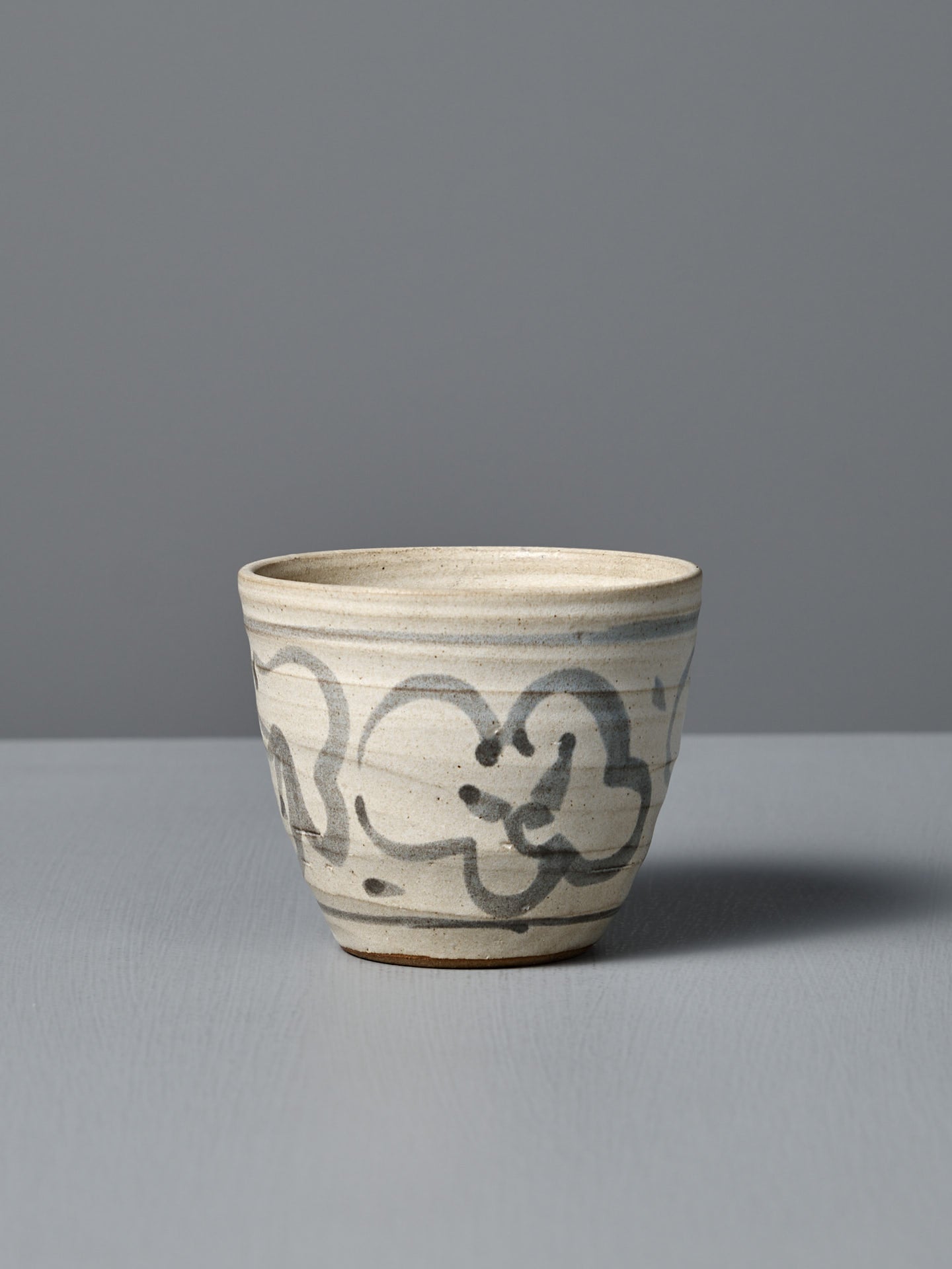 The Cup – Floral is a handmade ceramic cup featuring a beige glaze with dark abstract brushstrokes. It sits on a light gray surface against a gray background. Made in Aotearoa.