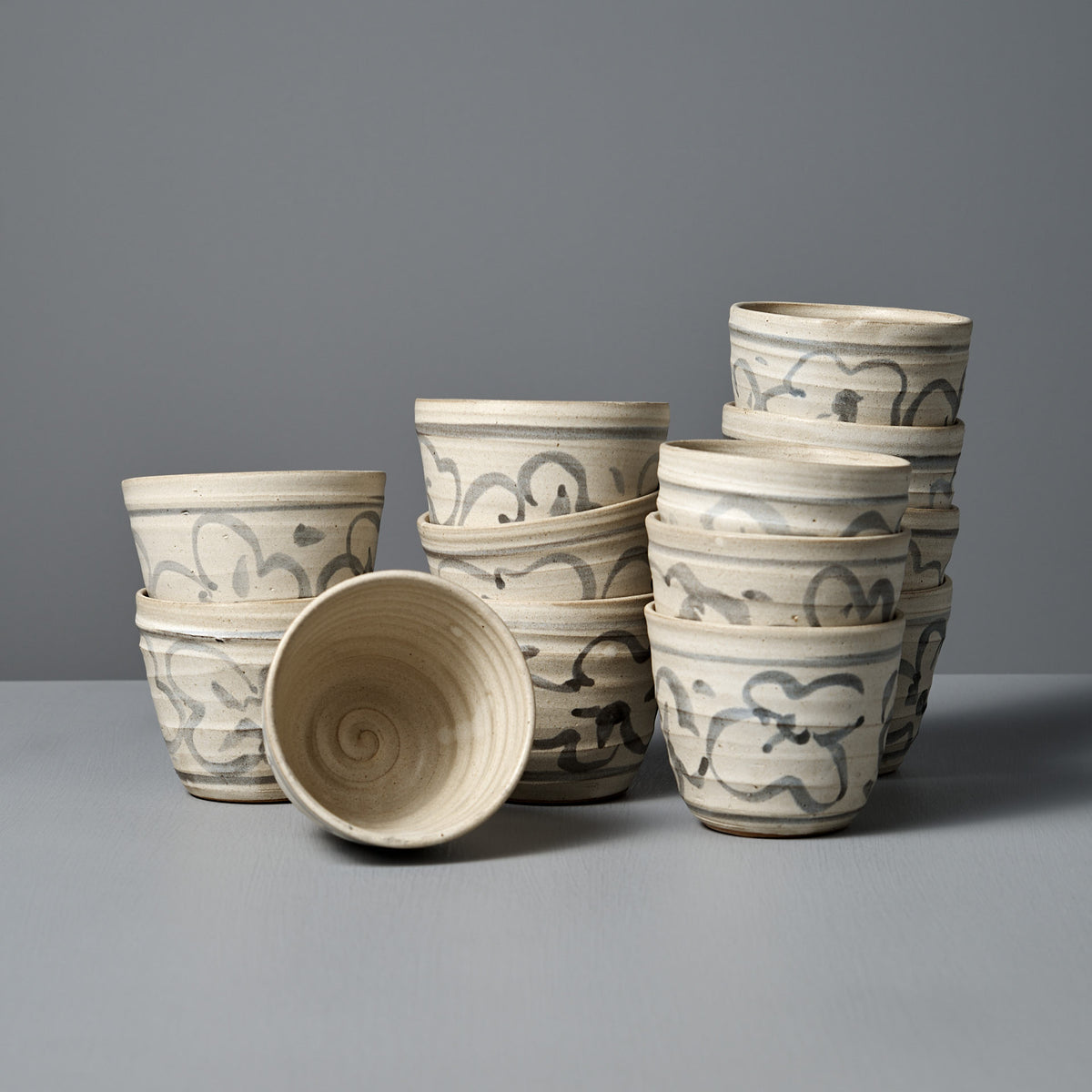 A collection of Cup – Floral handmade ceramic cups with gray and brown patterns are stacked in two uneven piles, while one cup with a matte cream glaze lies on its side in front.