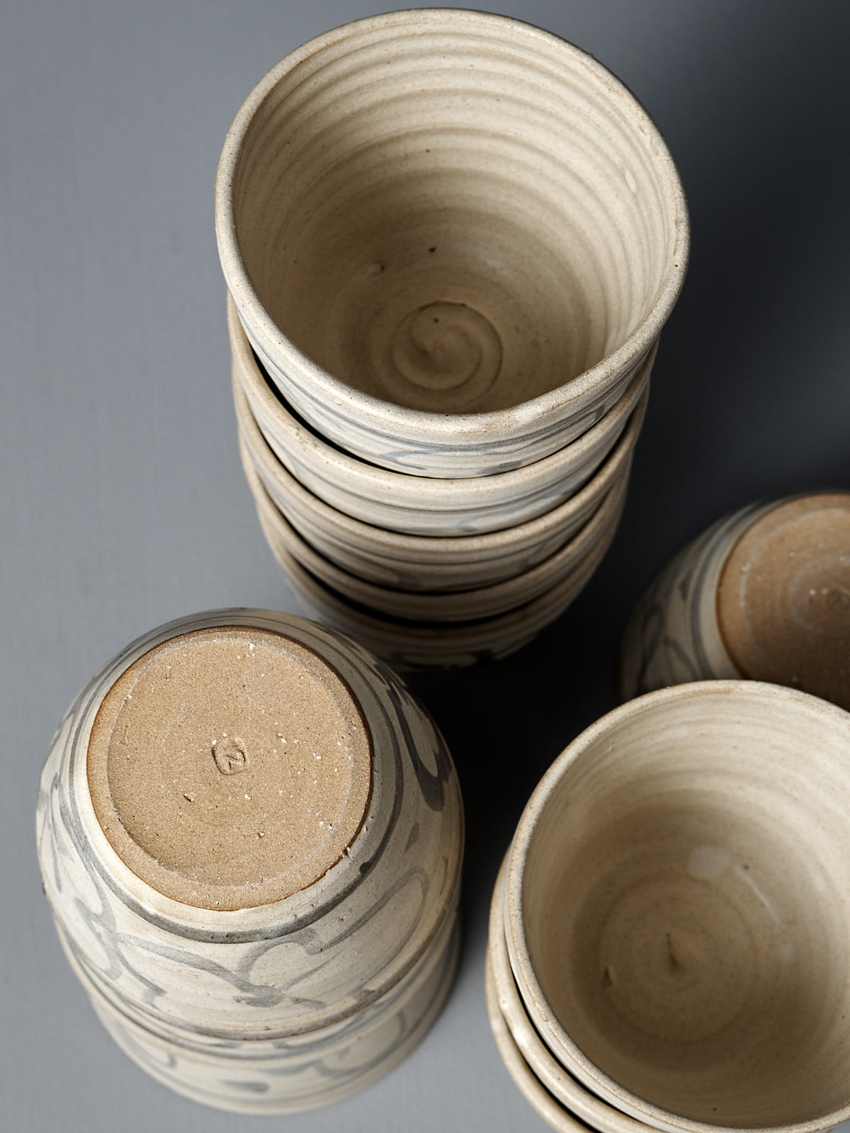 The Cup – Floral, a handmade ceramic cup, rests among stacked bowls on a gray surface, featuring textured patterns, natural clay tones, and a touch of matte cream glaze.