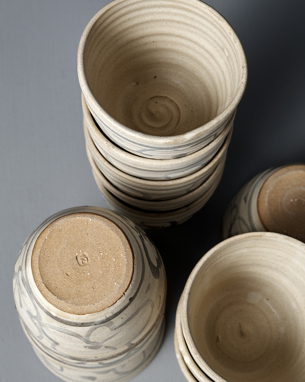 The Cup – Floral, a handmade ceramic cup, rests among stacked bowls on a gray surface, featuring textured patterns, natural clay tones, and a touch of matte cream glaze.