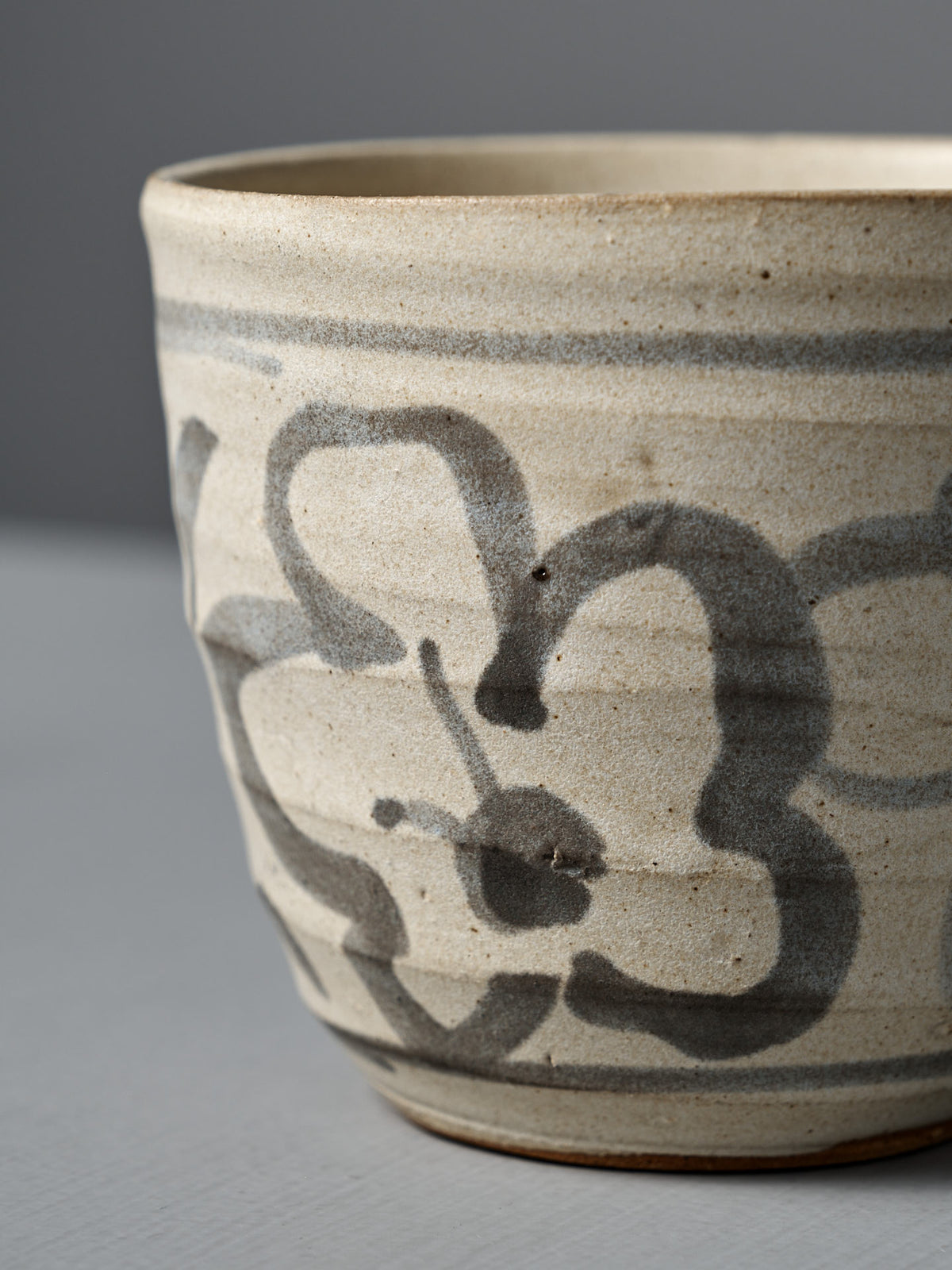 The Cup – Floral is a handmade ceramic cup featuring a matte cream glaze and hand-painted gray floral design, shown on a light gray surface with a neutral background.