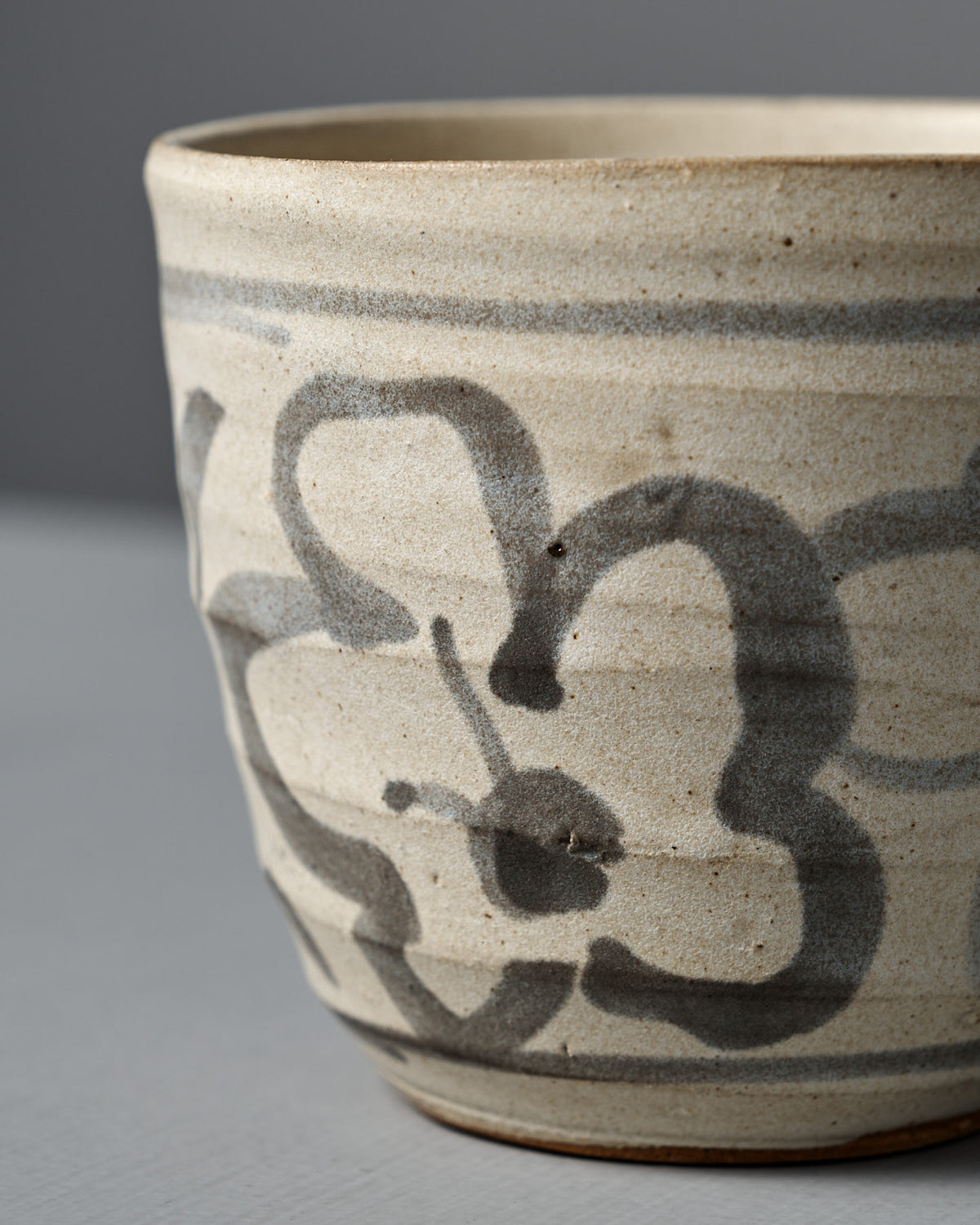 The Cup – Floral is a handmade ceramic cup featuring a matte cream glaze and hand-painted gray floral design, shown on a light gray surface with a neutral background.