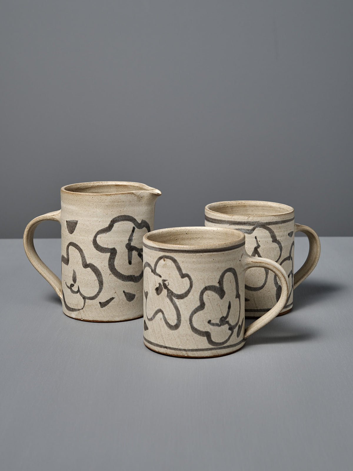 A Milk Jug – Floral featuring a beige wheel-thrown pottery design with black floral patterns is displayed on a gray surface against a plain gray background.