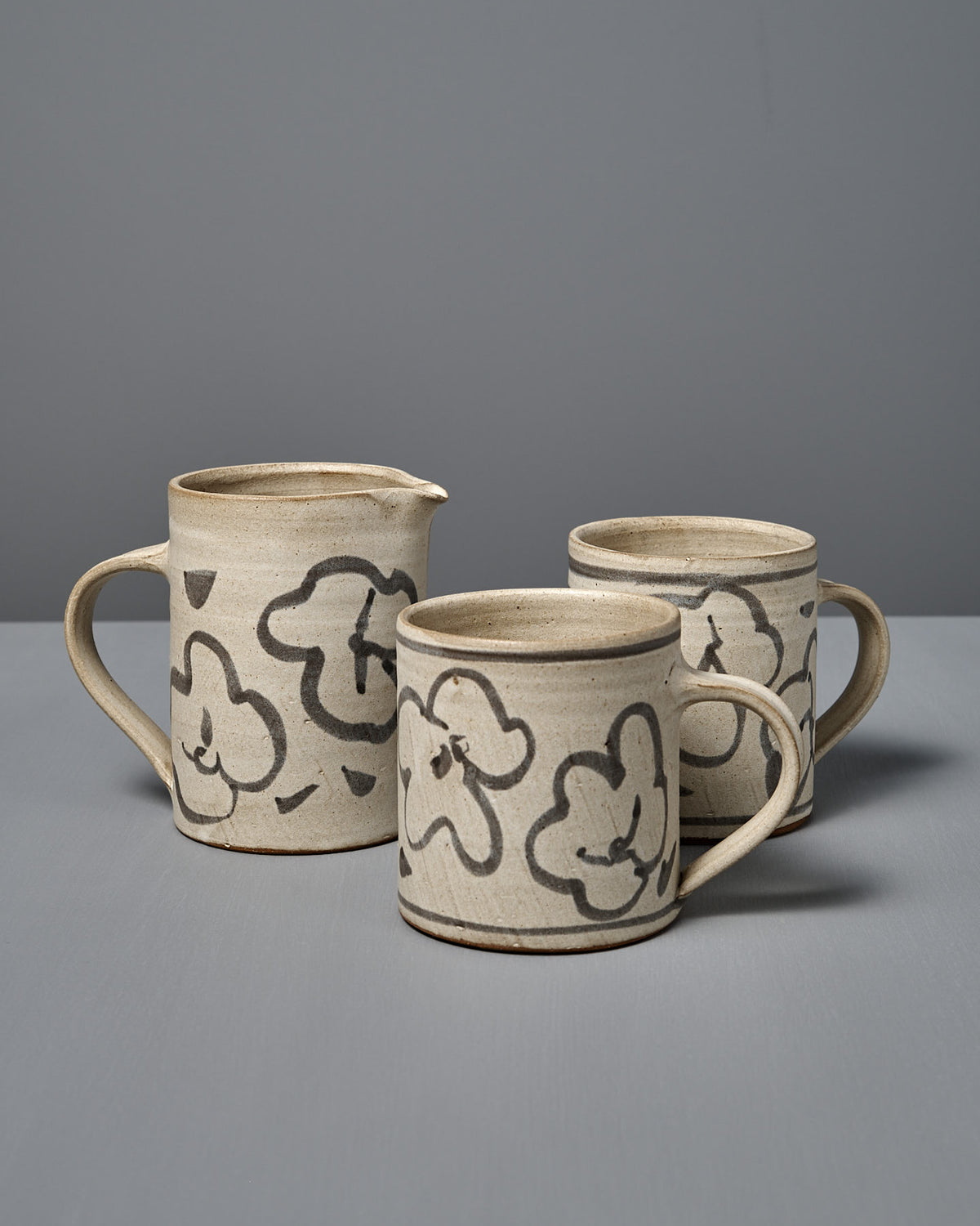 A Milk Jug – Floral featuring a beige wheel-thrown pottery design with black floral patterns is displayed on a gray surface against a plain gray background.