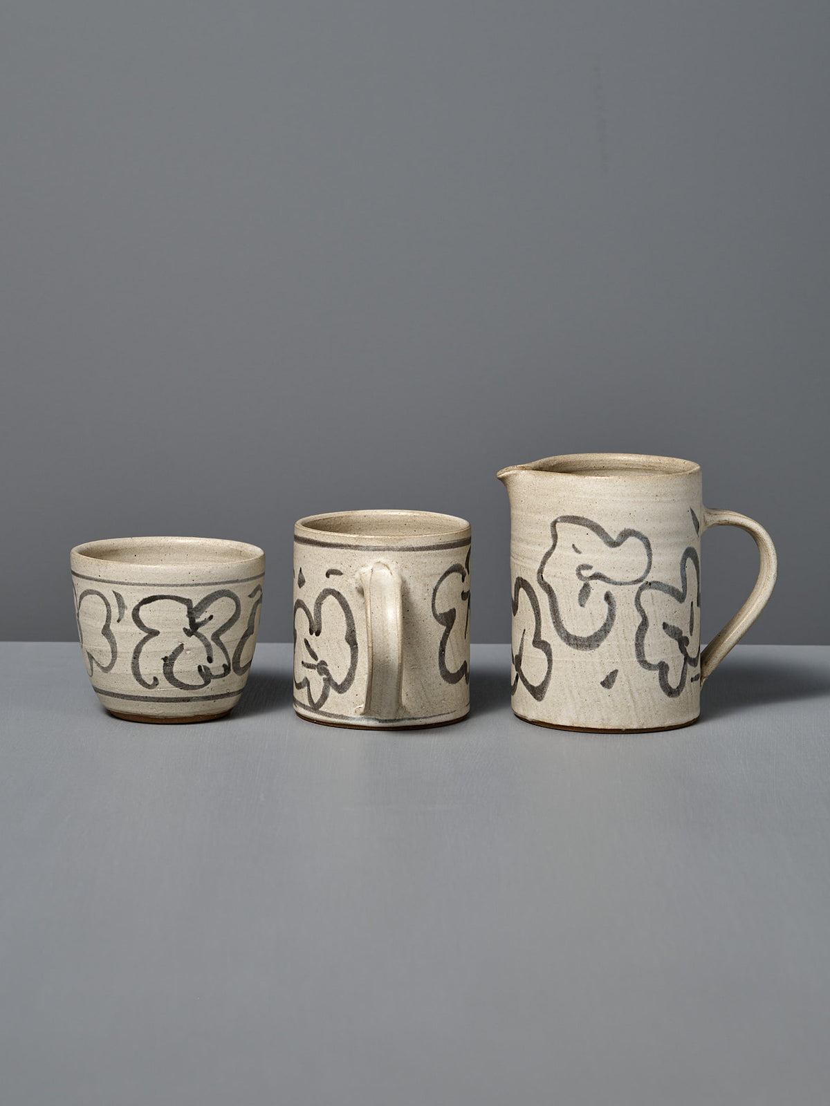 The Milk Jug – Floral, along with a cup and a mug, are displayed side by side on a gray surface, each wheel-thrown and adorned with black floral patterns over a white glaze.