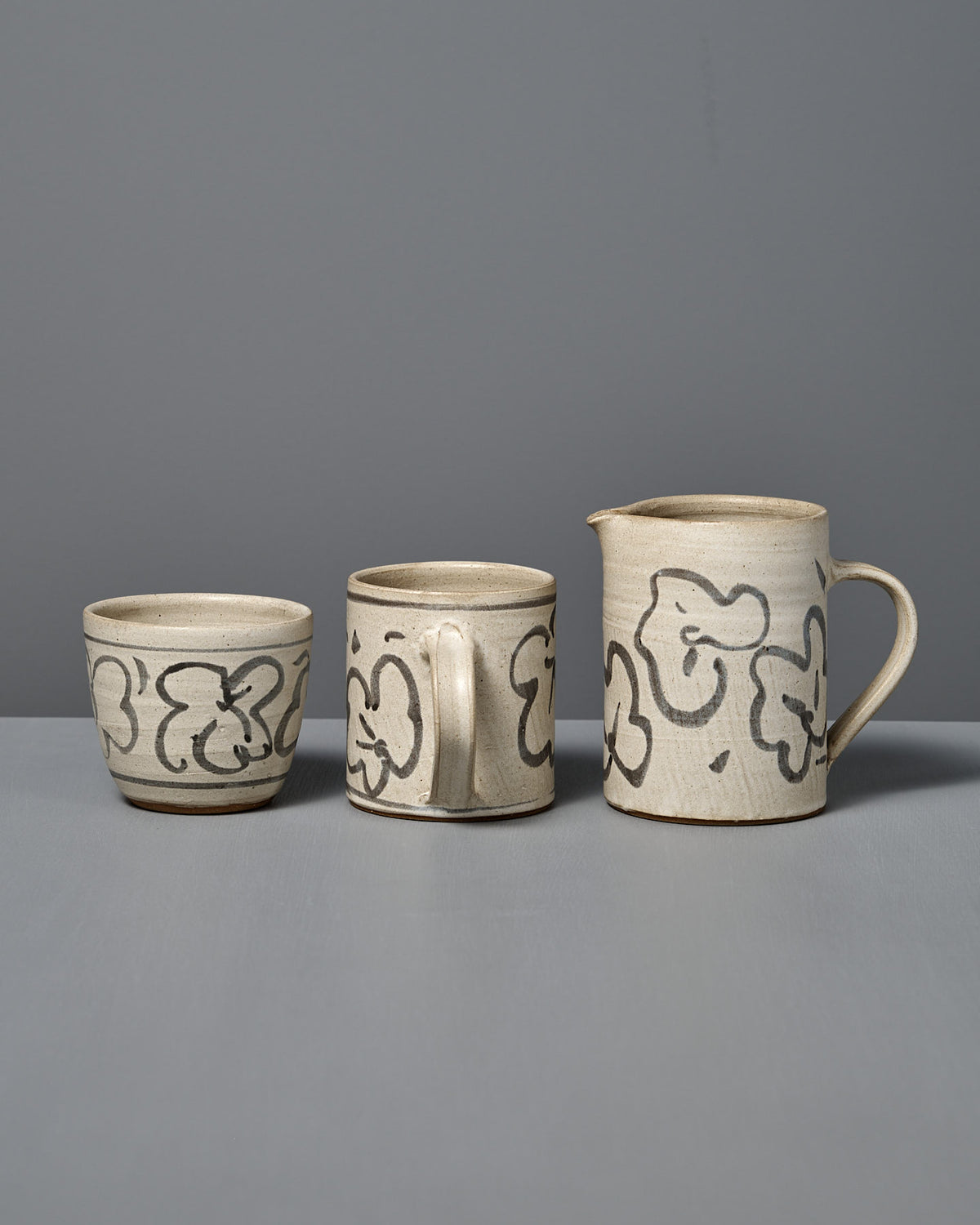 The Milk Jug – Floral, along with a cup and a mug, are displayed side by side on a gray surface, each wheel-thrown and adorned with black floral patterns over a white glaze.