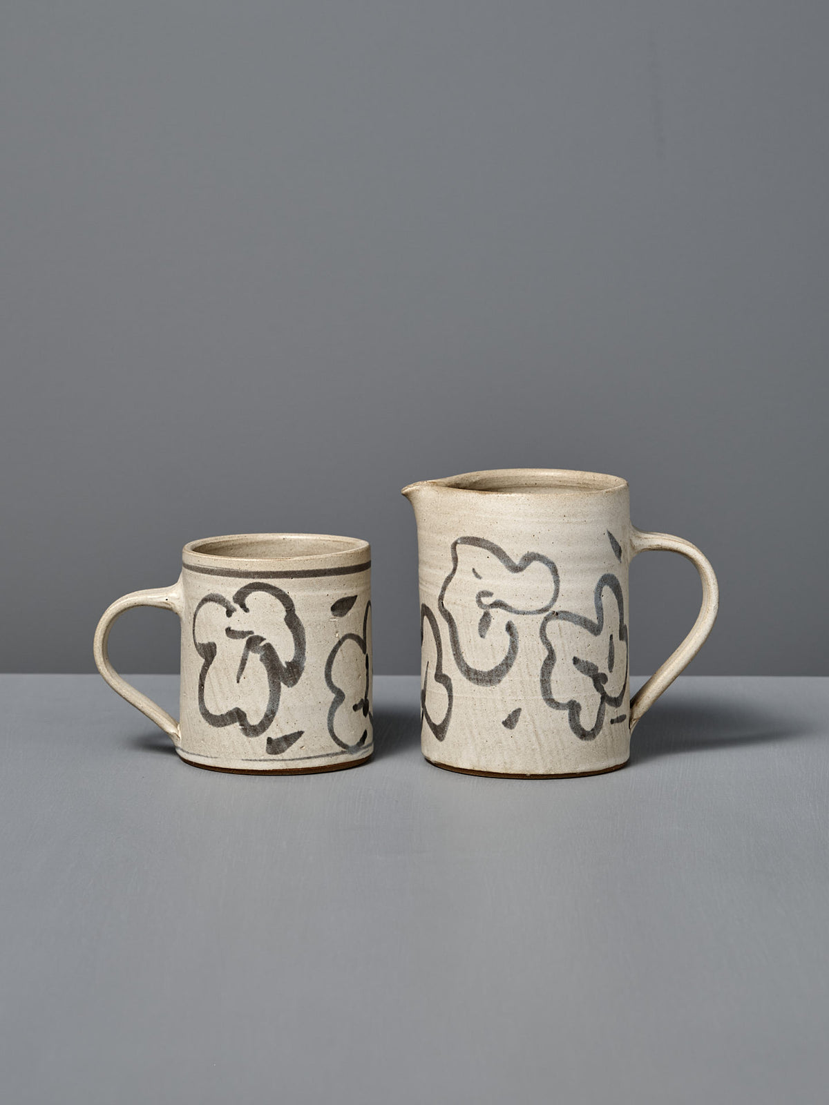 The Mug – Floral is a handmade ceramic mug featuring abstract black floral designs on a light background, displayed on a gray surface against a gray wall.