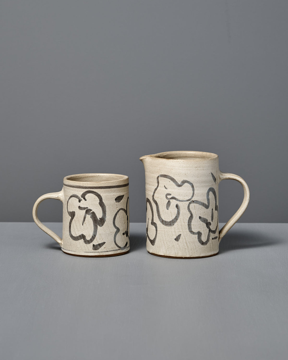 The Mug – Floral is a handmade ceramic mug featuring abstract black floral designs on a light background, displayed on a gray surface against a gray wall.