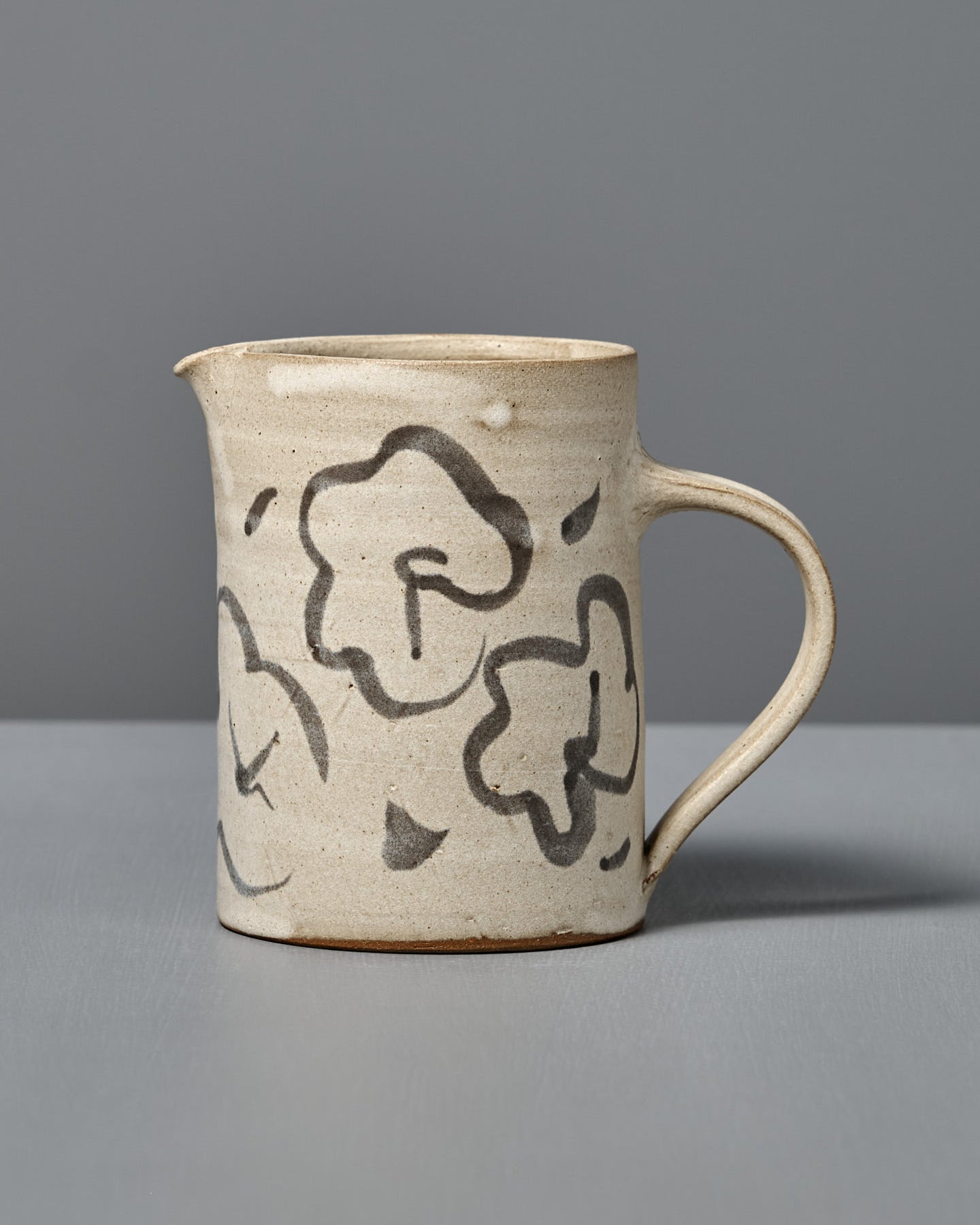 The Milk Jug – Floral features handmade beige ceramic with a handle and black floral designs, displayed on a gray surface and set against a gray background.