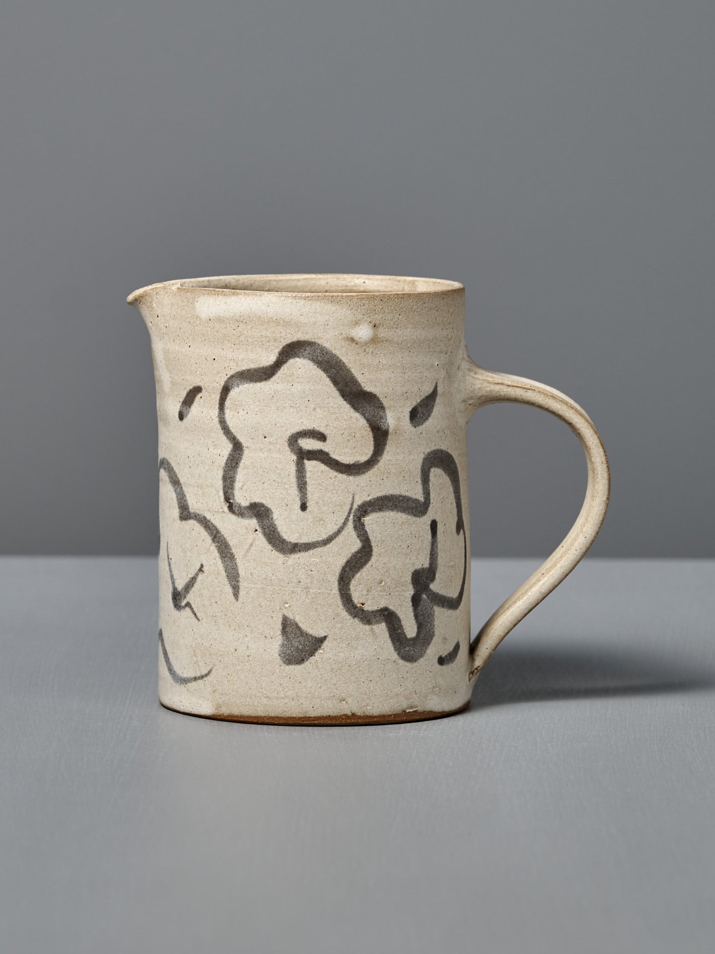 The Milk Jug – Floral features handmade beige ceramic with a handle and black floral designs, displayed on a gray surface and set against a gray background.