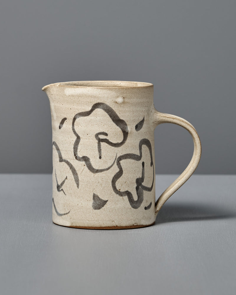 The Milk Jug – Floral features handmade beige ceramic with a handle and black floral designs, displayed on a gray surface and set against a gray background.