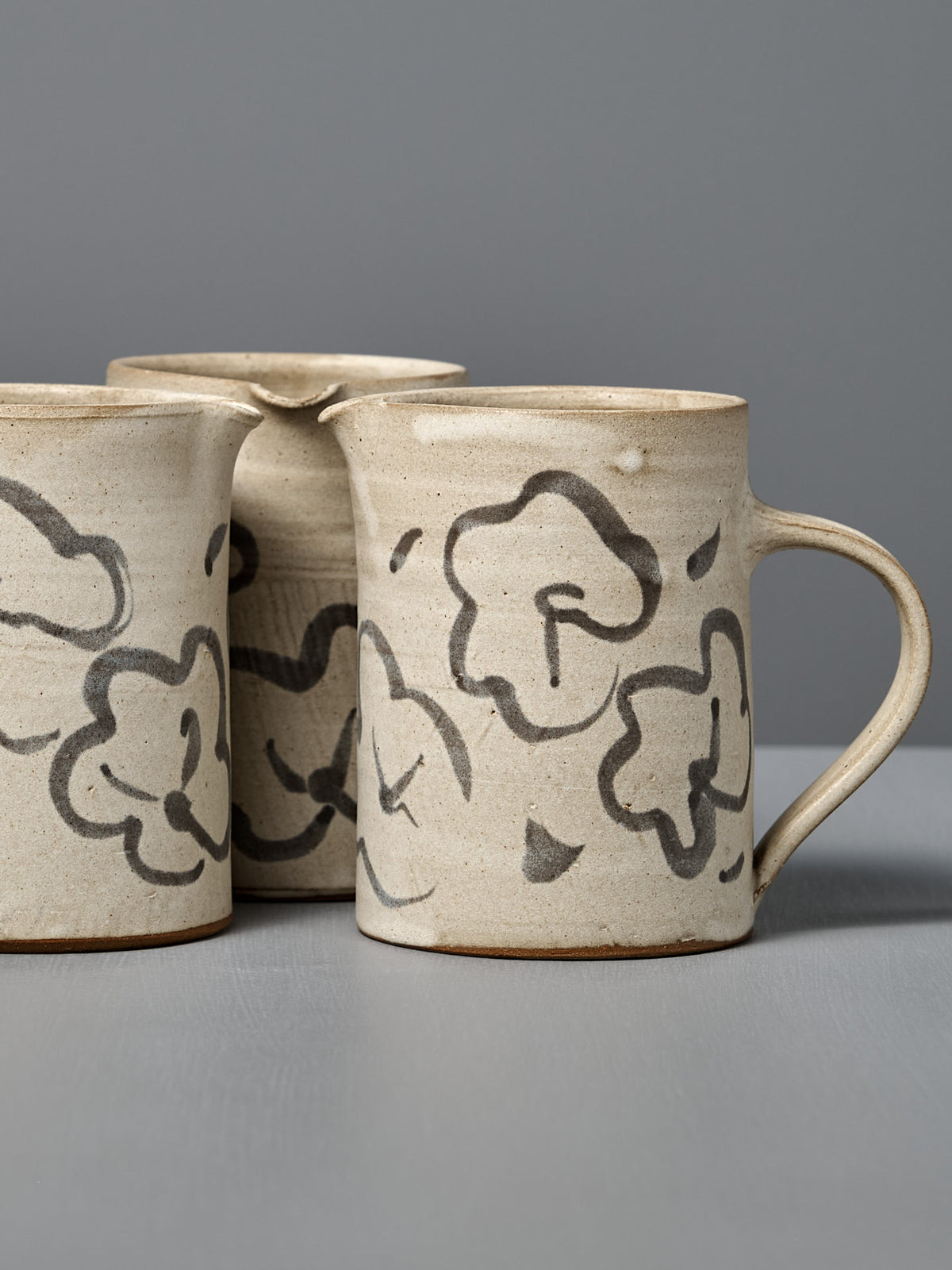 Three beige Milk Jug – Floral pieces with black abstract floral designs are arranged on a gray surface against a gray background.