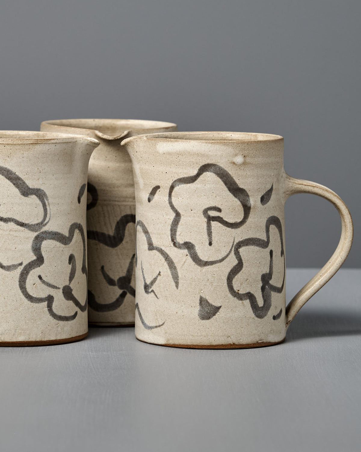 Three beige Milk Jug – Floral pieces with black abstract floral designs are arranged on a gray surface against a gray background.