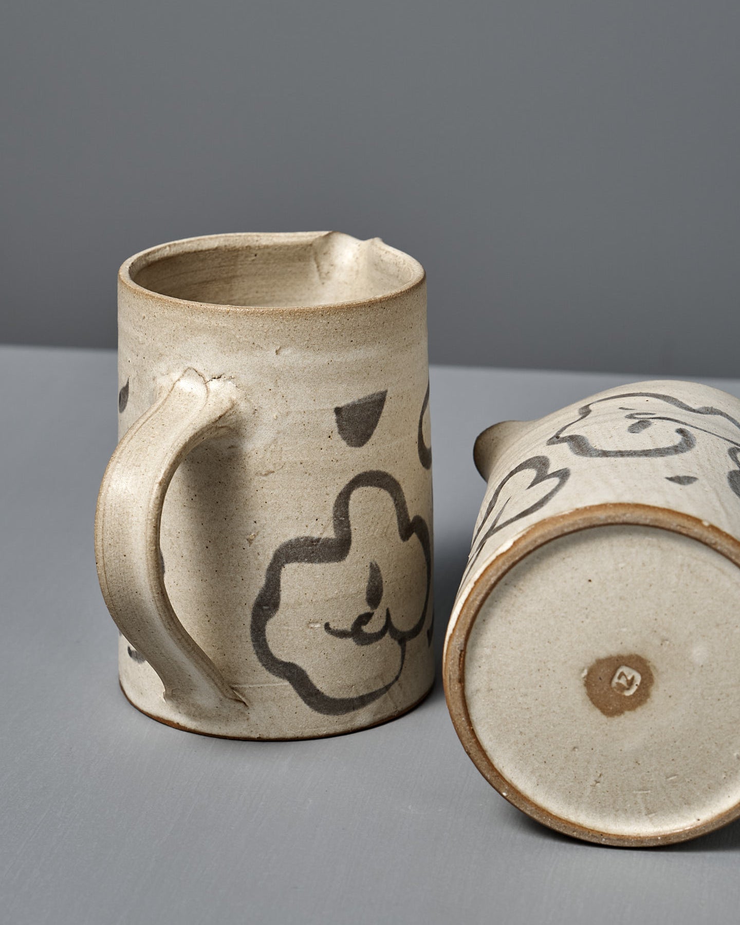 The Milk Jug – Floral features handmade beige ceramic with a handle and black floral designs, displayed on a gray surface and set against a gray background.