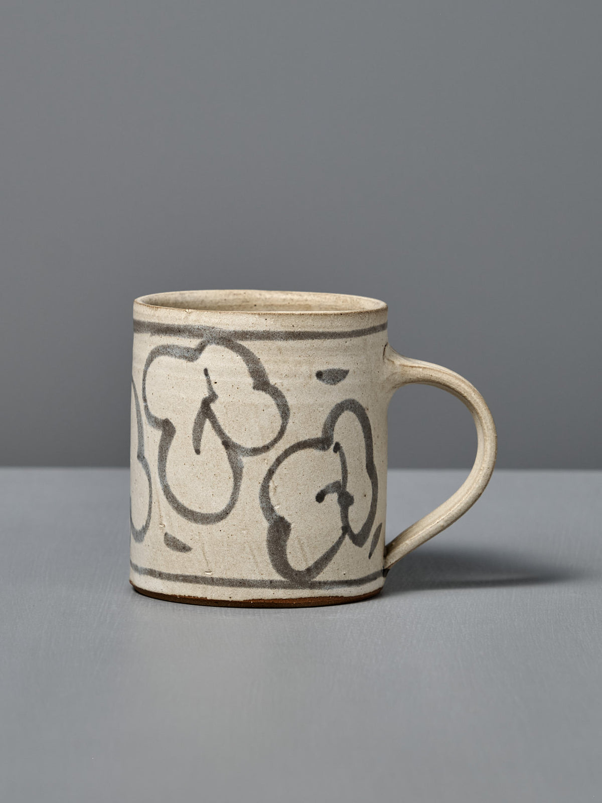 The Mug – Floral is a handmade ceramic mug with light beige glaze and abstract black flower line drawings, shown on a gray surface with a gray background.