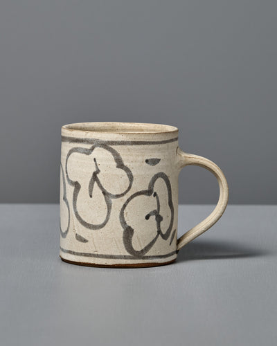 The Mug – Floral is a handmade ceramic mug with light beige glaze and abstract black flower line drawings, shown on a gray surface with a gray background.