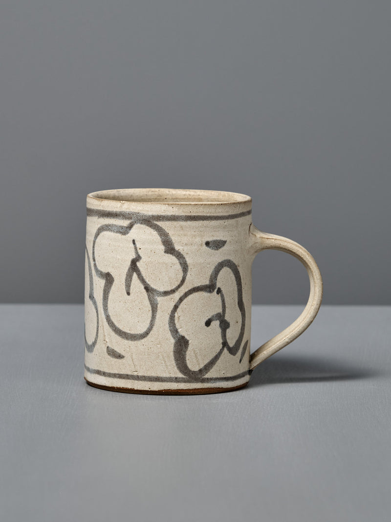 The Mug – Floral is a handmade ceramic mug with light beige glaze and abstract black flower line drawings, shown on a gray surface with a gray background.