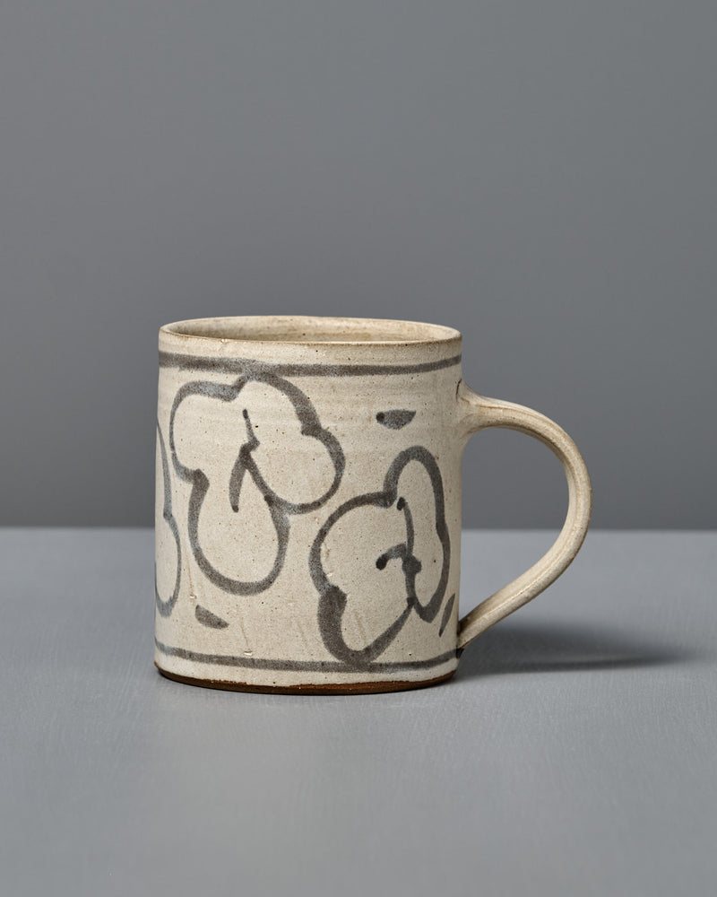 The Mug – Floral is a handmade ceramic mug with light beige glaze and abstract black flower line drawings, shown on a gray surface with a gray background.