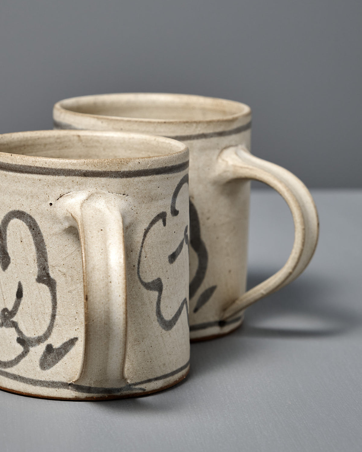 Two Mug – Floral handmade ceramic mugs featuring hand-painted abstract black designs are displayed on a gray surface with a gray background.