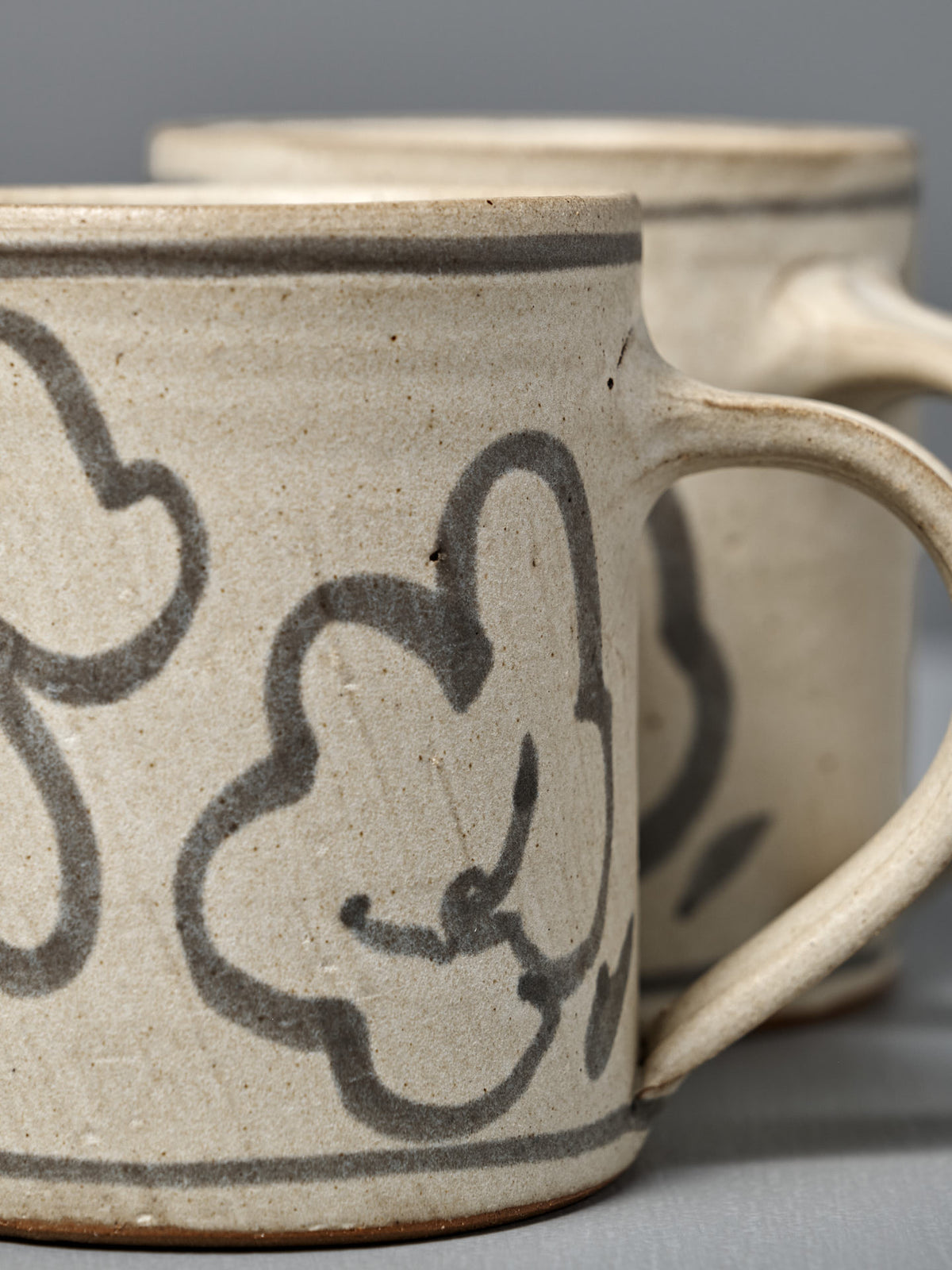 Two Mug – Floral ceramics in beige with gray floral designs sit side by side on a gray surface.