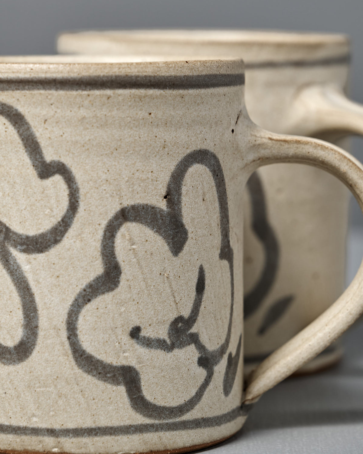 Two Mug – Floral ceramics in beige with gray floral designs sit side by side on a gray surface.