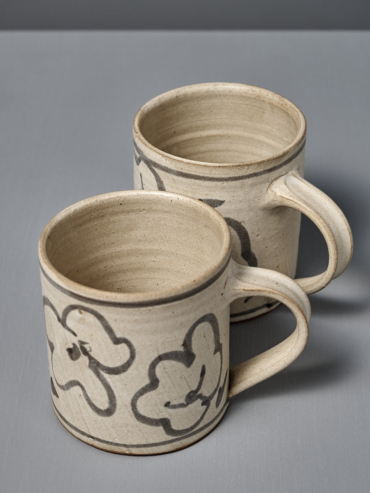 Two Mug – Floral pieces, wheel-thrown and featuring hand-painted abstract black line designs, are displayed on a gray surface.