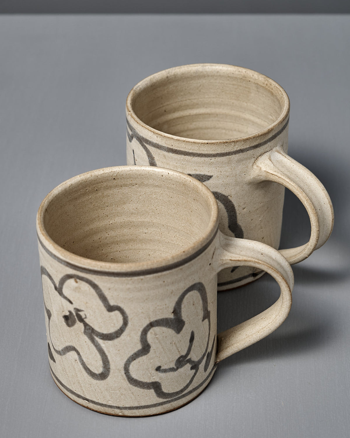 Two Mug – Floral pieces, wheel-thrown and featuring hand-painted abstract black line designs, are displayed on a gray surface.