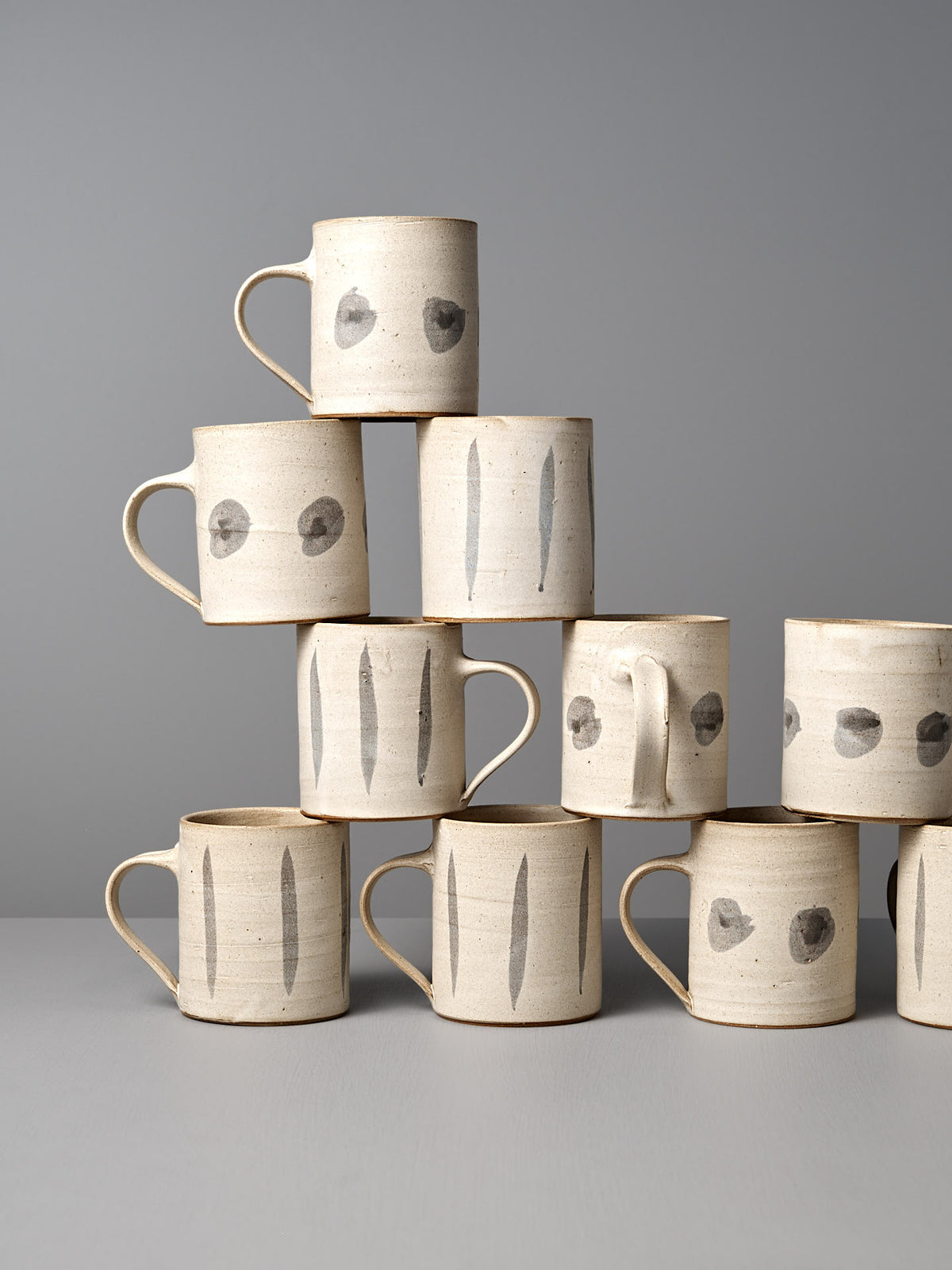 A pyramid stack of nine Zoë Isaacs Mug – Stripes, handmade from iron-rich clay with minimalist black stripe patterns on a gray surface, displayed against a gray background.