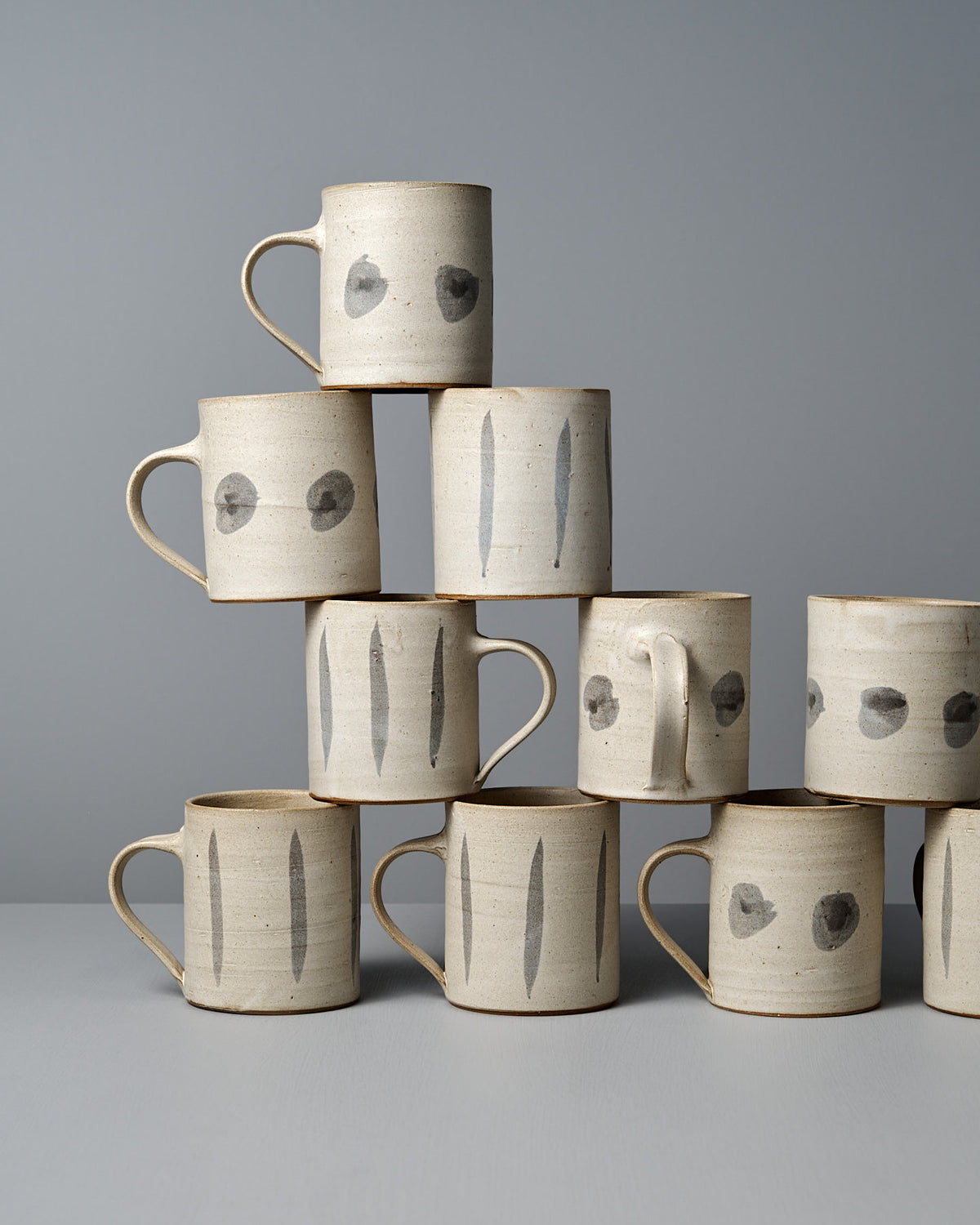 A pyramid stack of nine Zoë Isaacs Mug – Stripes, handmade from iron-rich clay with minimalist black stripe patterns on a gray surface, displayed against a gray background.