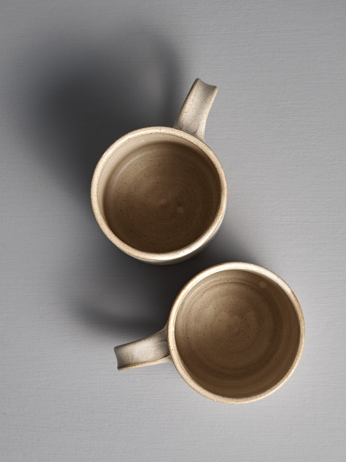 Two Zoë Isaacs Mug – Stripes ceramic mugs with handles, finished in matte cream glaze, are placed vertically and slightly overlapping on a gray surface, as seen from above.