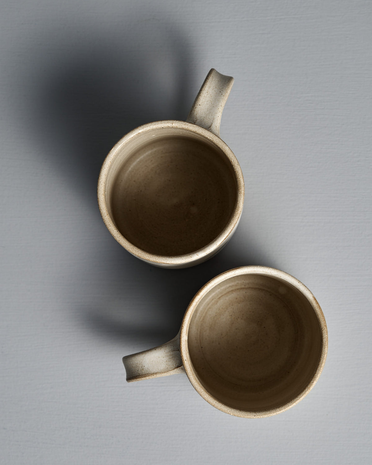 Two Zoë Isaacs Mug – Stripes ceramic mugs with handles, finished in matte cream glaze, are placed vertically and slightly overlapping on a gray surface, as seen from above.