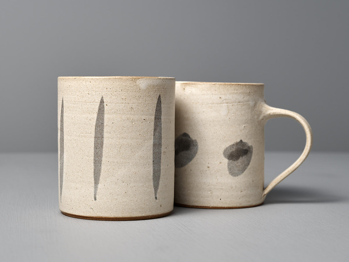 The Mug – Stripes by Zoë Isaacs is a handmade cup crafted from iron-rich clay, showcasing simple gray brushstroke designs and a matte cream glaze, displayed on a light gray surface with a neutral background.