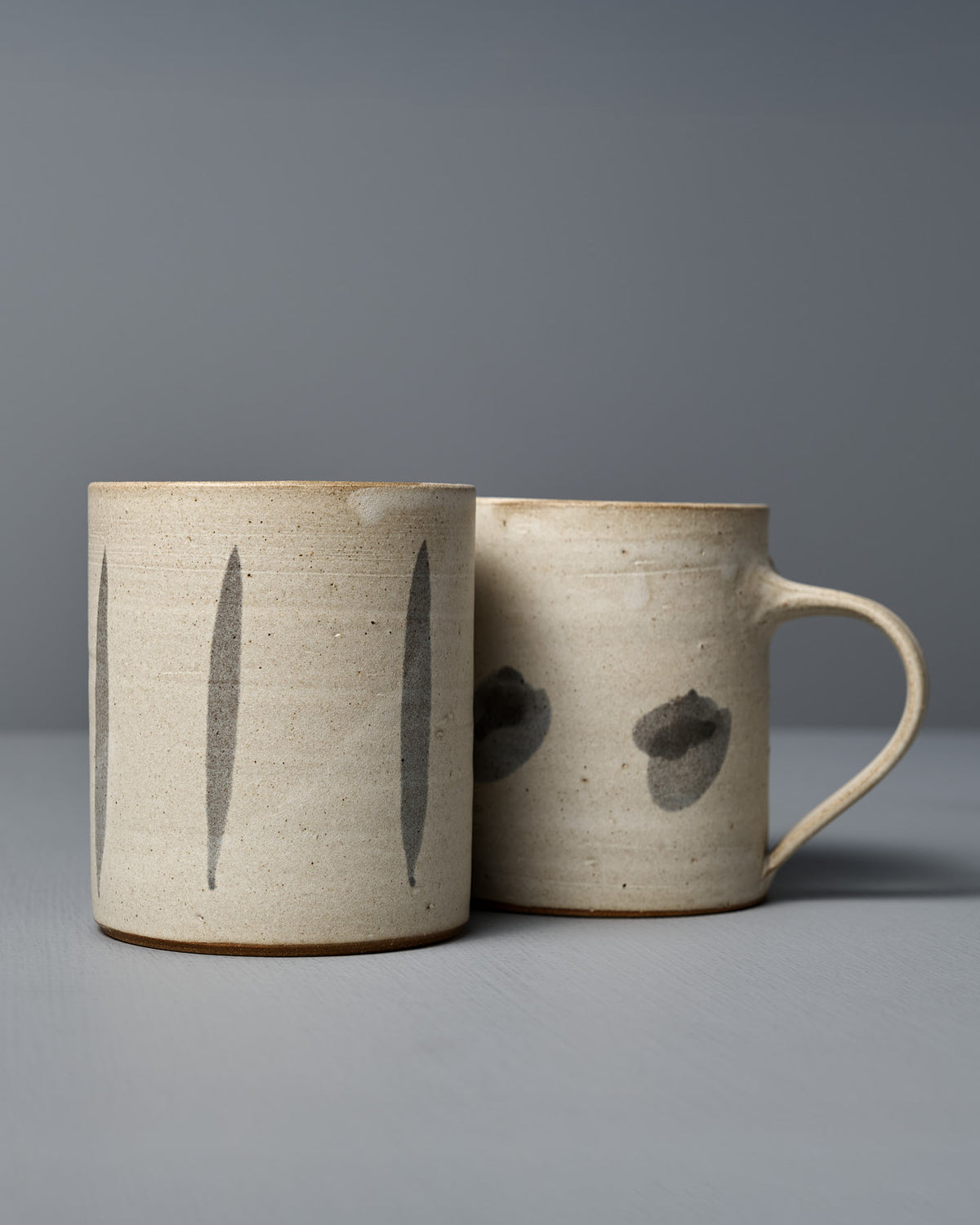 The Mug – Stripes by Zoë Isaacs is a handmade cup crafted from iron-rich clay, showcasing simple gray brushstroke designs and a matte cream glaze, displayed on a light gray surface with a neutral background.