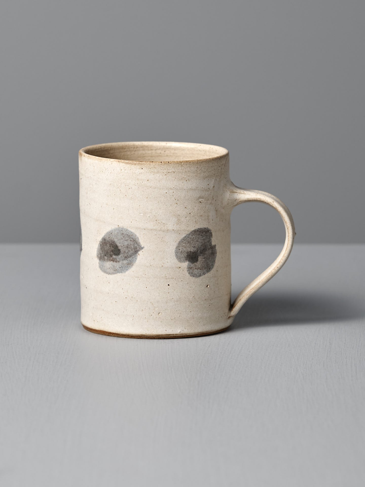 The Mug – Spots by Zoë Isaacs is a handmade ceramic mug with a matte cream glaze, curved handle, and two dark gray circular spots on its light beige surface, shown resting on a gray surface against a gray background.