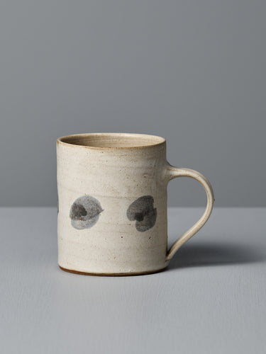 The Mug – Spots by Zoë Isaacs is a handmade ceramic mug with a matte cream glaze, curved handle, and two dark gray circular spots on its light beige surface, shown resting on a gray surface against a gray background.