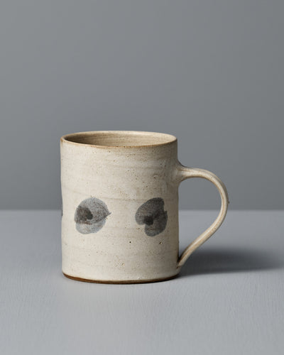 The Mug – Spots by Zoë Isaacs is a handmade ceramic mug with a matte cream glaze, curved handle, and two dark gray circular spots on its light beige surface, shown resting on a gray surface against a gray background.