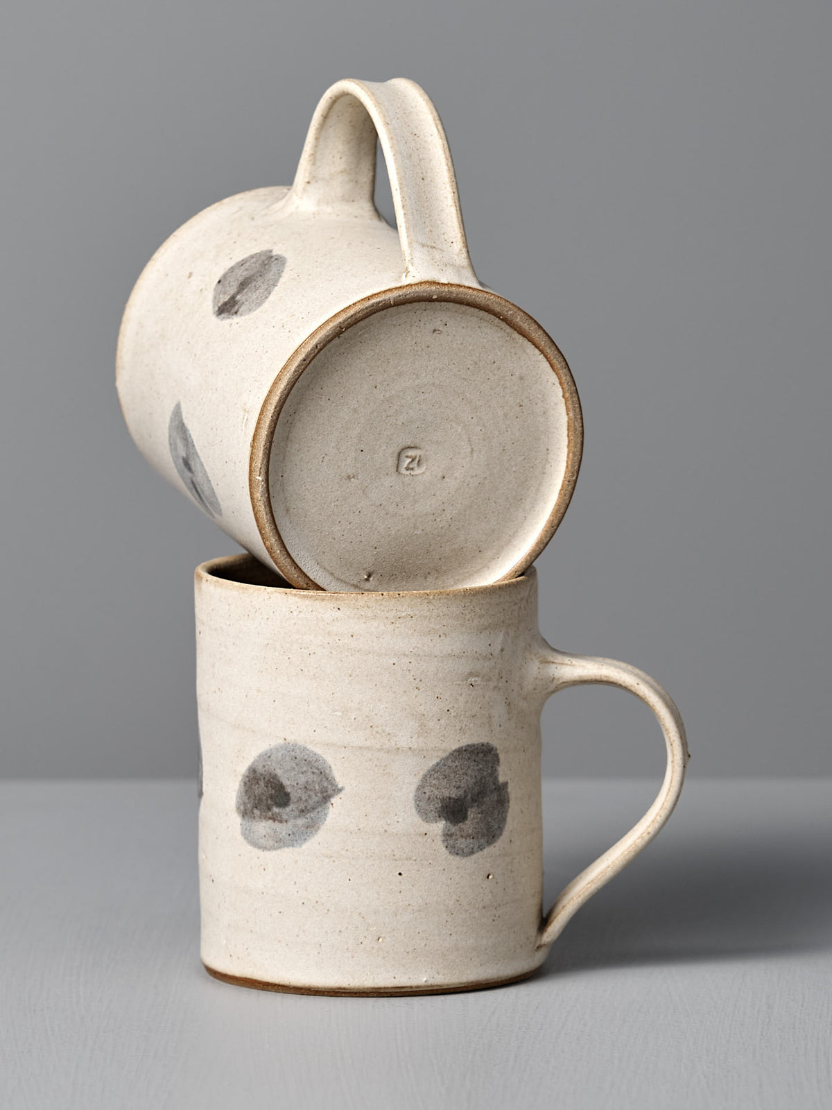 Two Zoë Isaacs &quot;Mug – Spots&quot; handmade cups crafted from wheel-thrown clay, featuring a matte cream glaze with gray spots—one mug rests sideways atop the other against a plain gray background.