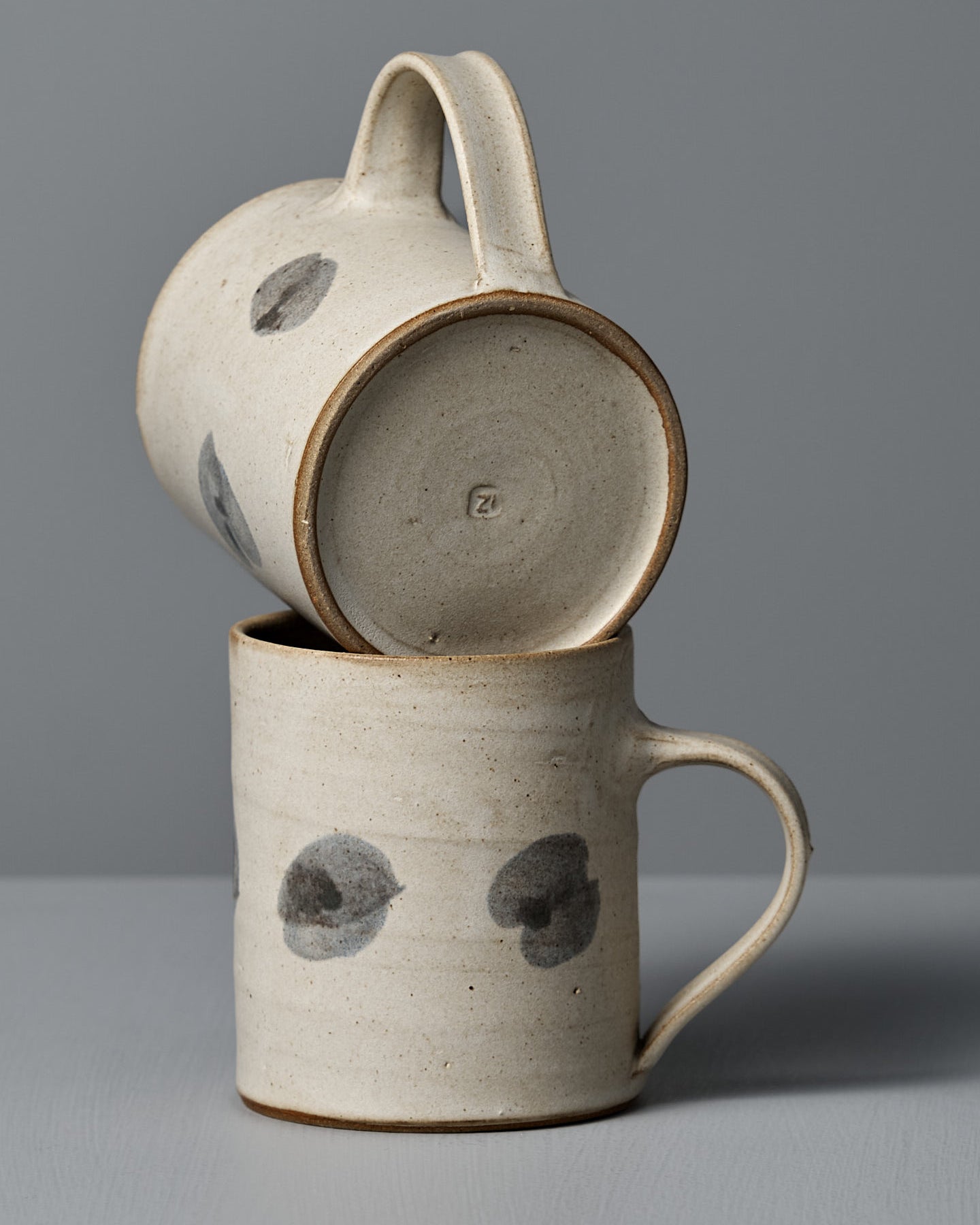 The Mug – Spots by Zoë Isaacs is a handmade ceramic mug with a matte cream glaze, curved handle, and two dark gray circular spots on its light beige surface, shown resting on a gray surface against a gray background.