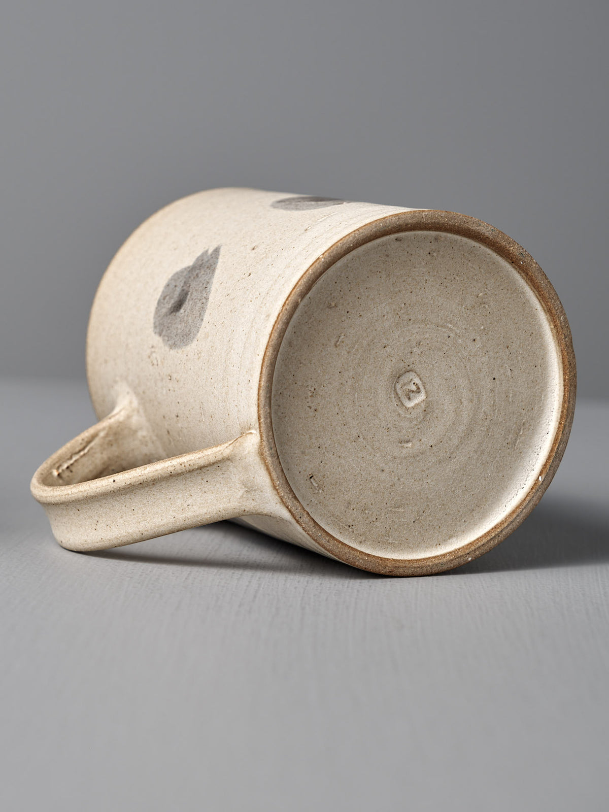 The Mug – Spots by Zoë Isaacs is a handmade cup with a simple, speckled matte cream glaze and a small imprint on the base, shown lying on its side on a gray surface.