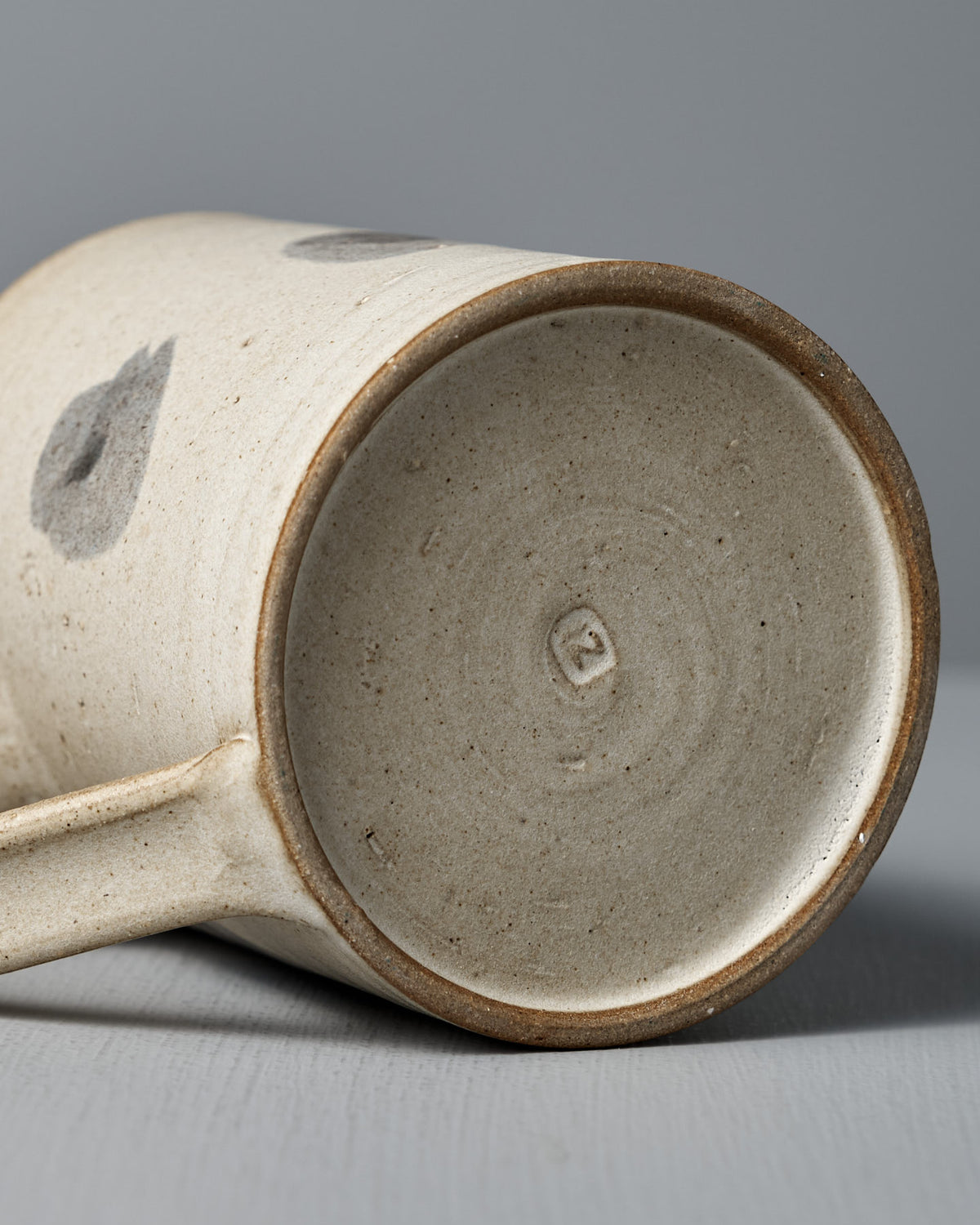 The Mug – Spots by Zoë Isaacs is a handmade cup with a simple, speckled matte cream glaze and a small imprint on the base, shown lying on its side on a gray surface.