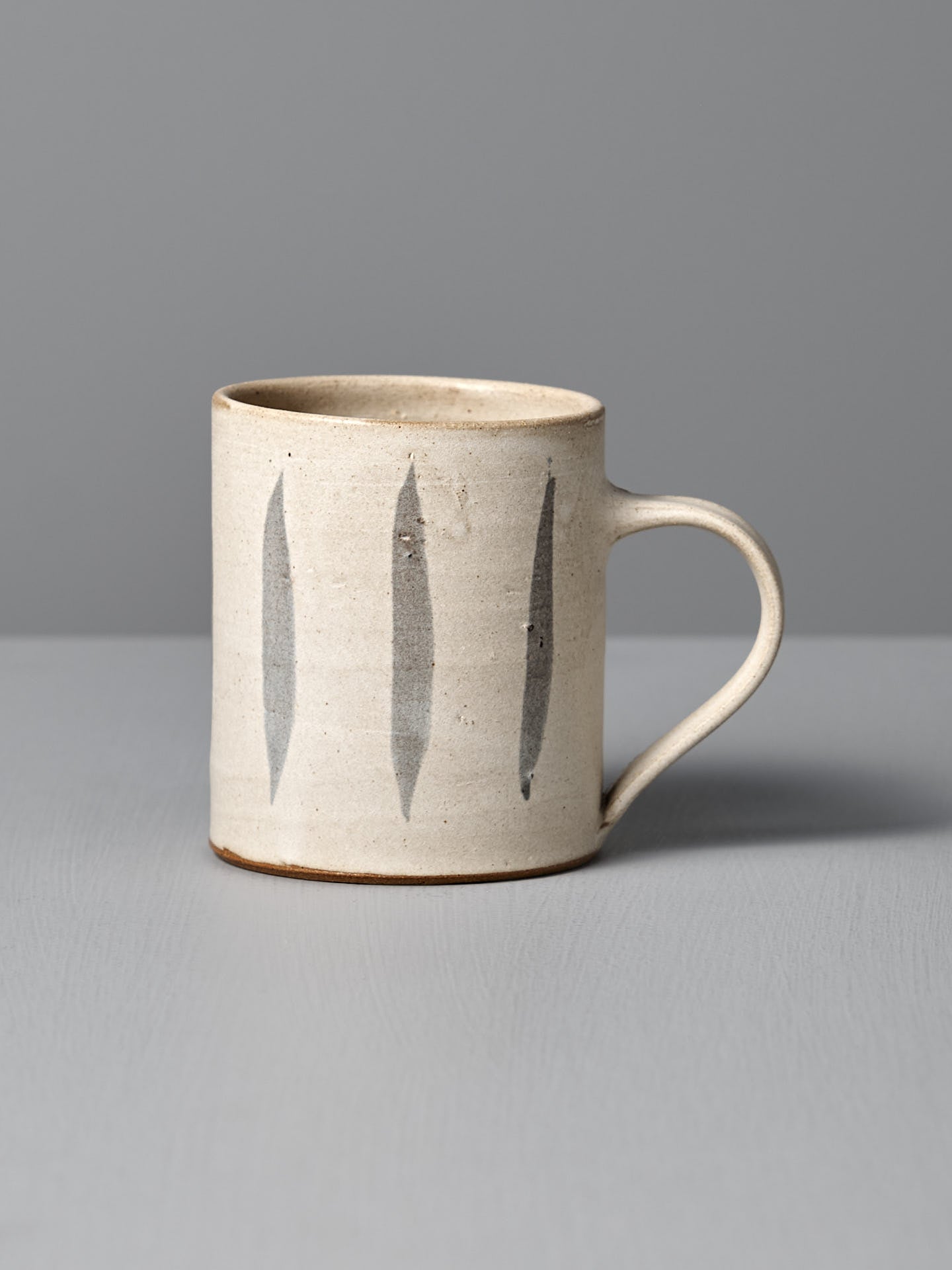 The Zoë Isaacs Mug – Stripes is a handmade beige ceramic mug with a handle, featuring three vertical dark gray stripes and a matte cream glaze, shown on a light gray surface with a gray background.