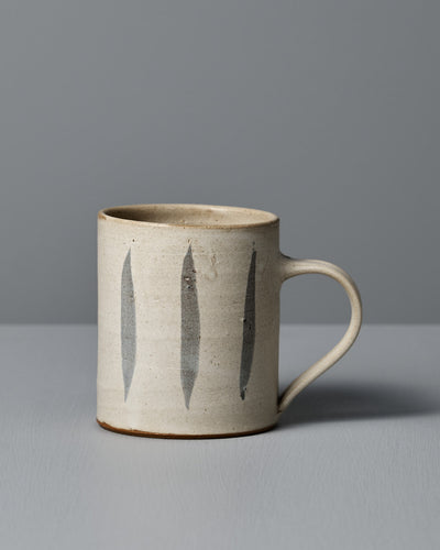 The Zoë Isaacs Mug – Stripes is a handmade beige ceramic mug with a handle, featuring three vertical dark gray stripes and a matte cream glaze, shown on a light gray surface with a gray background.