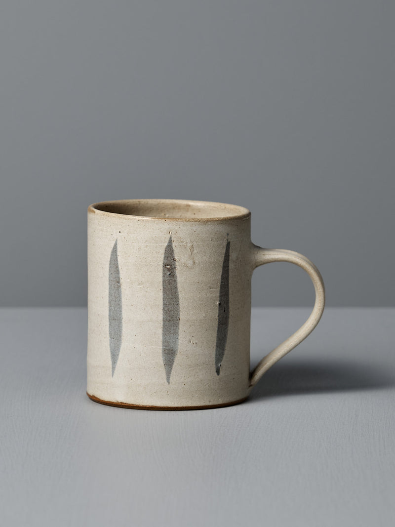 The Zoë Isaacs Mug – Stripes is a handmade beige ceramic mug with a handle, featuring three vertical dark gray stripes and a matte cream glaze, shown on a light gray surface with a gray background.