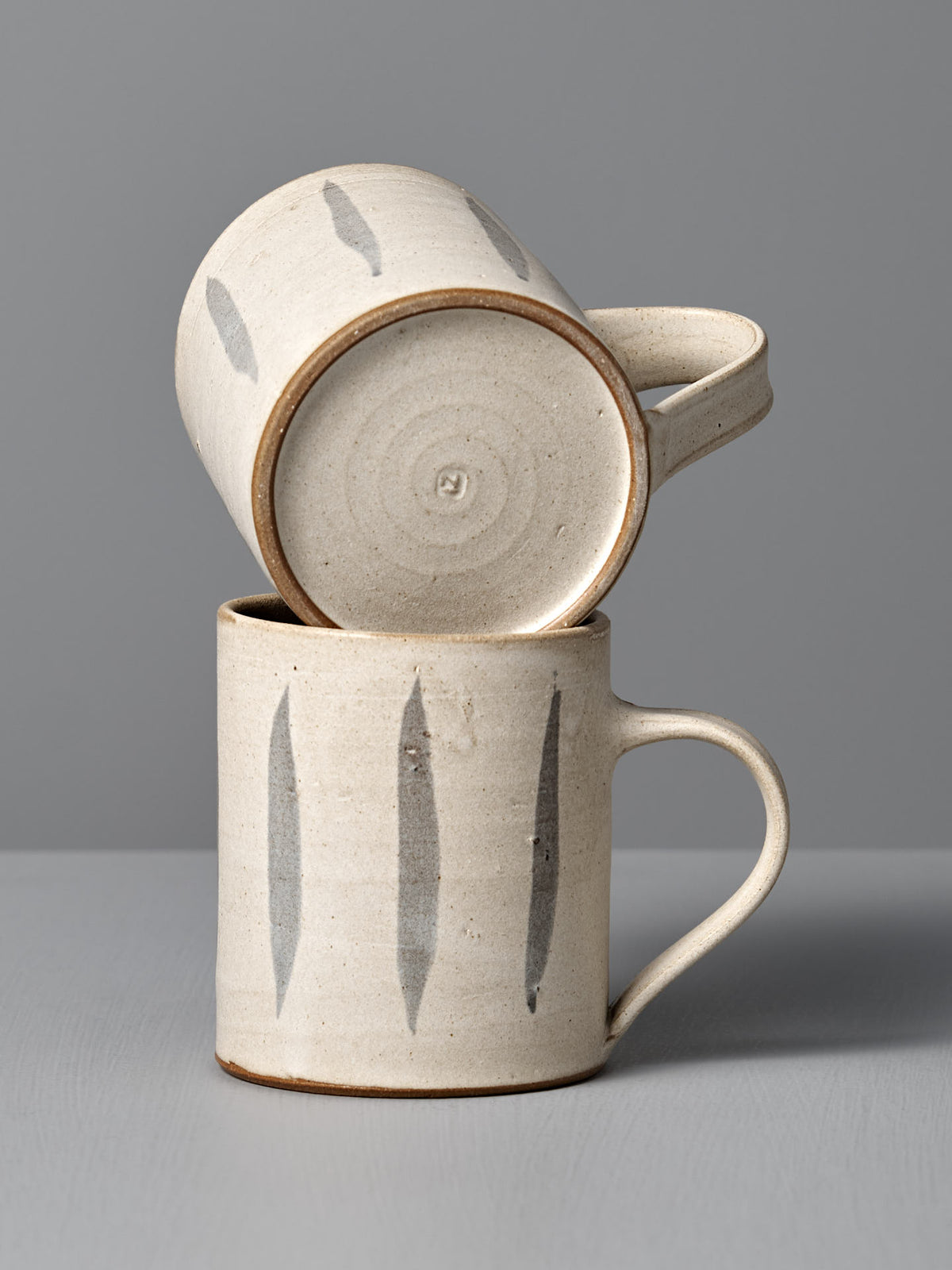Two Zoë Isaacs Mug – Stripes ceramic mugs, featuring vertical gray brushstroke designs and crafted from iron-rich clay, are stacked—one resting horizontally atop the other—against a plain gray background.