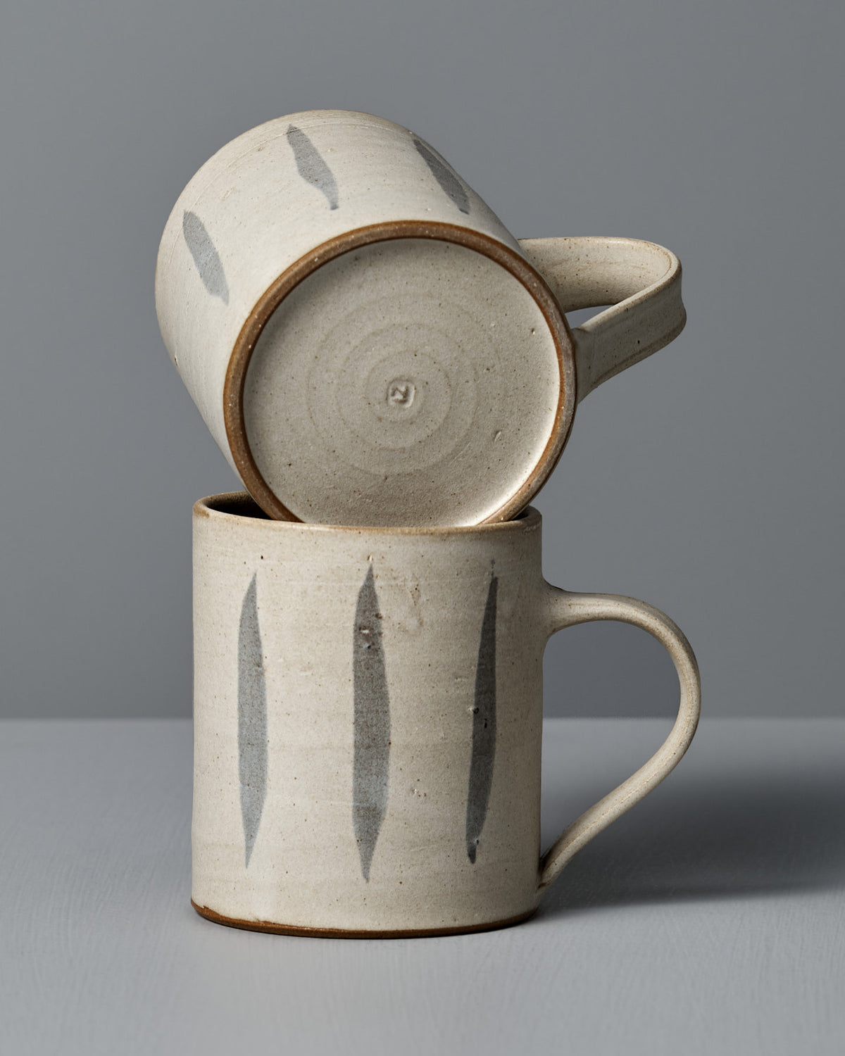 Two Zoë Isaacs Mug – Stripes ceramic mugs, featuring vertical gray brushstroke designs and crafted from iron-rich clay, are stacked—one resting horizontally atop the other—against a plain gray background.