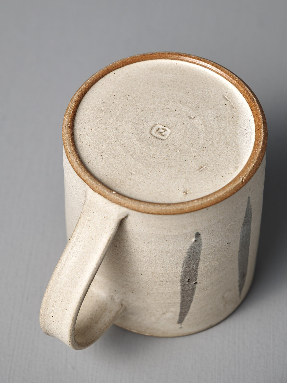 The Mug – Stripes by Zoë Isaacs is a handmade ceramic mug featuring two vertical brushstrokes, a matte cream glaze, and a circular stamp on its unglazed, iron-rich clay base.